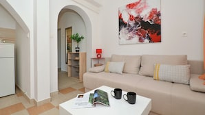 Un salon moderne avec un canapé beige, une table basse sur laquelle reposent un magazine et deux tasses noires, une lampe rouge et un grand tableau abstrait accroché au mur.