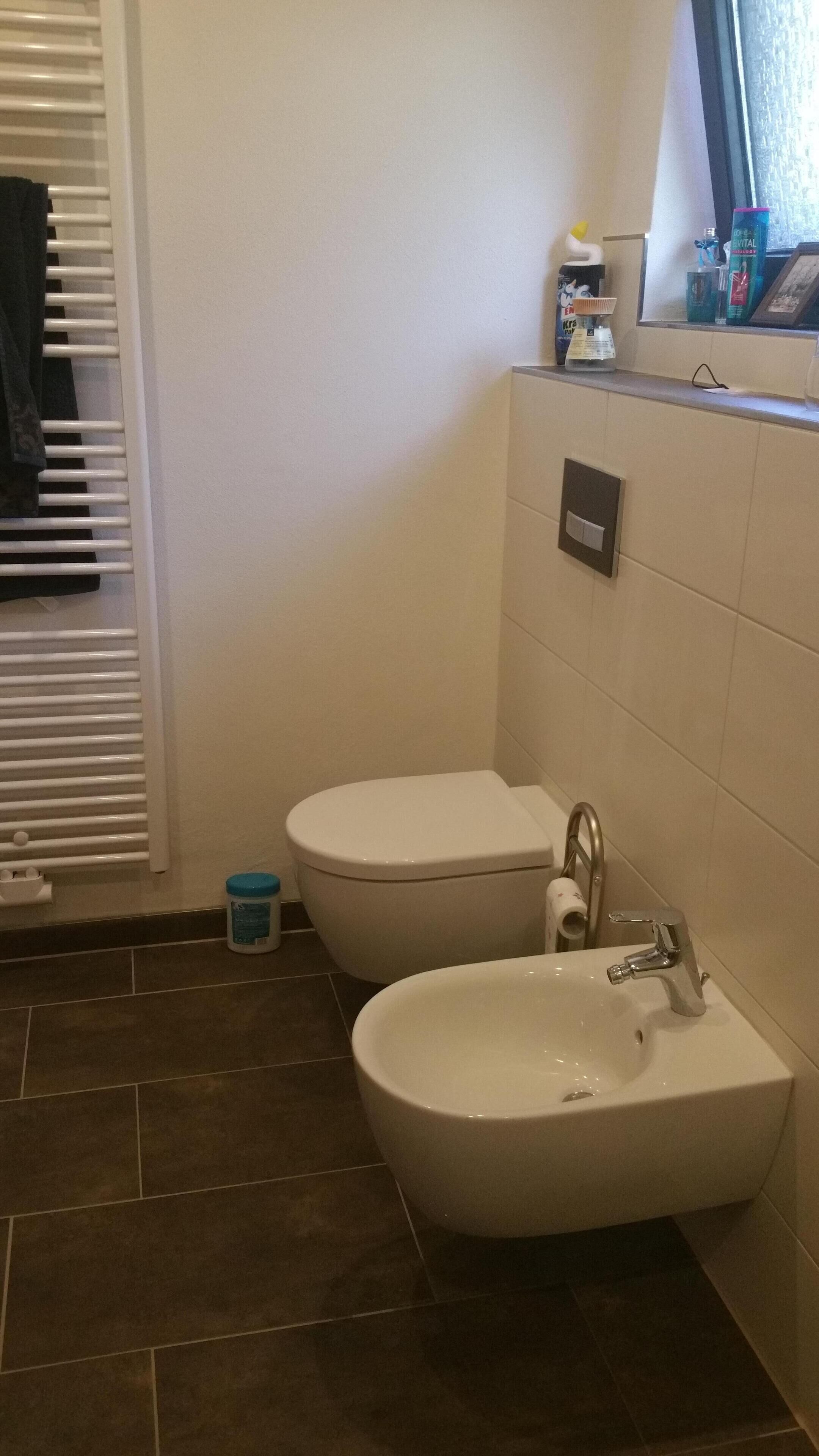 Un baño con inodoro, bidé y lavamanos. Hay un perchero con toallas, un pequeño cubo de basura y artículos de aseo en la encimera.