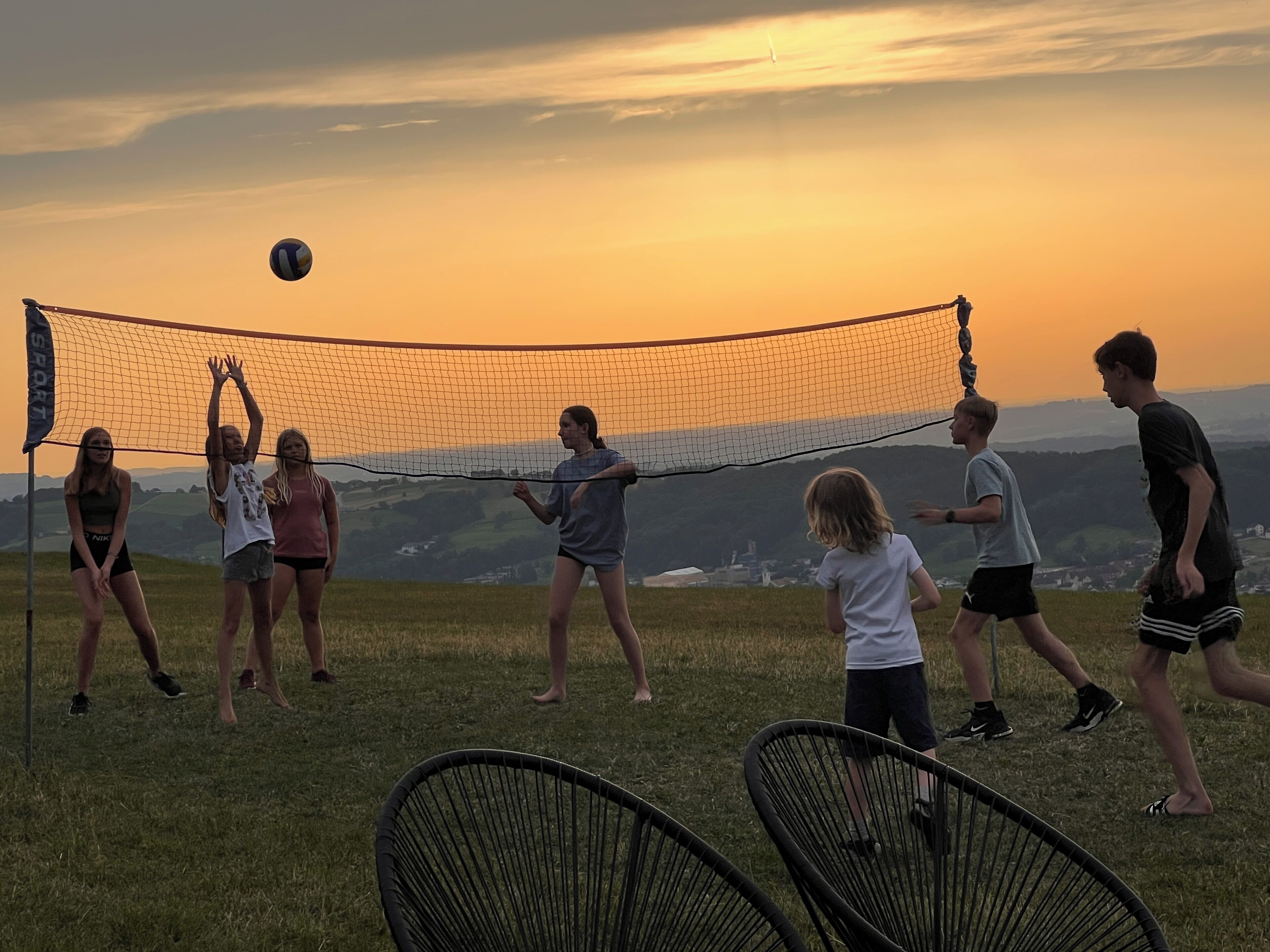 Un groupe de personnes joue au volley-ball sur un terrain herbeux, avec un filet, des chaises et une vue panoramique sur des collines et des bâtiments en arrière-plan.
