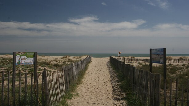 Ein Sandweg am Strand mit Holzzäunen und Schildern, der zum Meer führt.