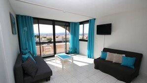 Ein modernes Wohnzimmer mit einer schwarzen Couch, einem weißen Couchtisch und einem weiteren schwarzen Sofa mit blauen Kissen. Ein großes Fenster mit blauen Vorhängen, ein flachbildfernseher an der Wand und Blick auf Gebäude sowie einen klaren Himmel.