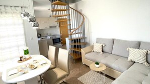 A modern living room with a grey sofa, a dining table with chairs, a spiral staircase, and a kitchen area.