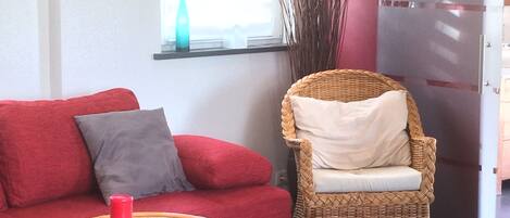 A red sofa with a grey pillow, a wicker chair with a white cushion, a small round table with a red candle and magazines, and a window with a bottle on the sill.
