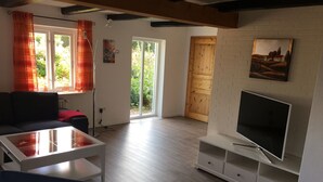 Ein Wohnzimmer mit einer Couch, einem Glastisch, einem Fernseher auf einem weißen Schrank und einer Holztür, die in einen anderen Raum führt.