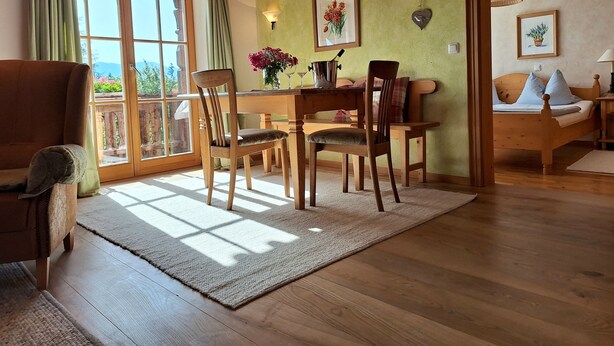 A cozy room with a wooden dining table, chairs, and a sofa. There is a large window with curtains, a wall-mounted lamp, and a framed picture. - Heissbauer Vacation Rental "Rosentraum" with Mountain Views and Two Bedrooms (Wackersberg)