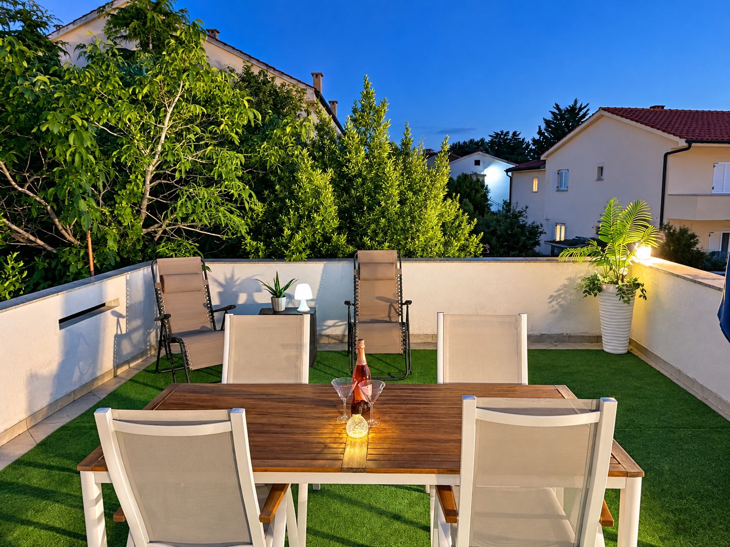 An outdoor patio with a wooden table and chairs, surrounded by a white wall and greenery.