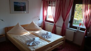 Een net opgemaakt bed met kussens en handdoeken, een ingelijst schilderij aan de muur, een raam met gordijnen en een radiator.