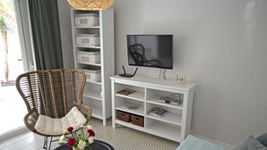 Een woonkamer met een rieten stoel, een zwarte tafel met een bloevas, een televisie op een witte kast, en opbergplanken.