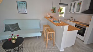 Een ruimtelijk ingerichte leefruimte met kitchenette, een lichtblauwe bank, een eettafeltje en een barstoel.