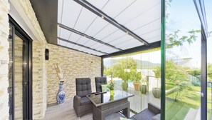 A modern outdoor patio with wicker furniture, a glass table, and a stone wall. - Apartment with pool on the Cote Azur (Bagnols en foret)