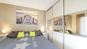A bedroom with a bed, a nightstand, a lamp, a wall-mounted painting, and a large mirror. - Apartment with pool on the Cote Azur (Bagnols en foret)