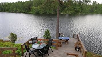 A wooden deck with a round table and chairs, overlooking a calm lake with a forested shoreline.