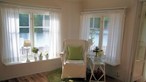 A room with a white chair, a small table with a lamp, and a window with curtains.