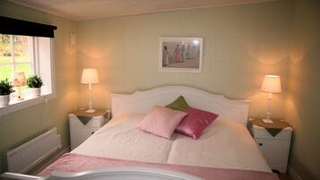 A neatly arranged bedroom with a bed, two bedside lamps, a radiator, and a window with potted plants.