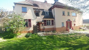 Ein zweigeschossiges Haus mit geneigtem Dach, einer kleinen Veranda und einem Garten mit Rasenfläche und Baum.