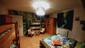 Ein Schlafzimmer mit einem Holzbettgestell, einem Einzelbett mit farbenfroher Bettdecke im Schmetterlingsdesign, einem Sessel, einem kleinen Tisch, einem großen Gemälde eines Wasserfalls und einem Wandgemälde mit Waldmotiv.