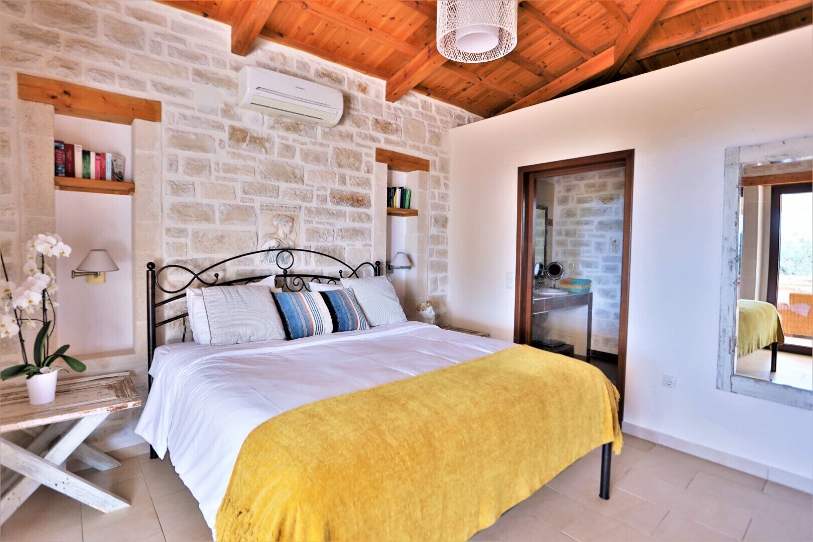 A bedroom with a stone wall, wooden ceiling beams, a bed with white bedding and a yellow throw, a bedside table with a lamp, and a window with a view.