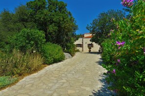 Un chemin de pierre mène à un bâtiment surmonté d’un toit en tuiles rouges, qui est entouré d’une végétation luxuriante et de plantes en fleurs.