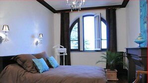 Une chambre à coucher avec un grand lit, une table de chevet, une chaise, une fenêtre avec des rideaux et un lustre.