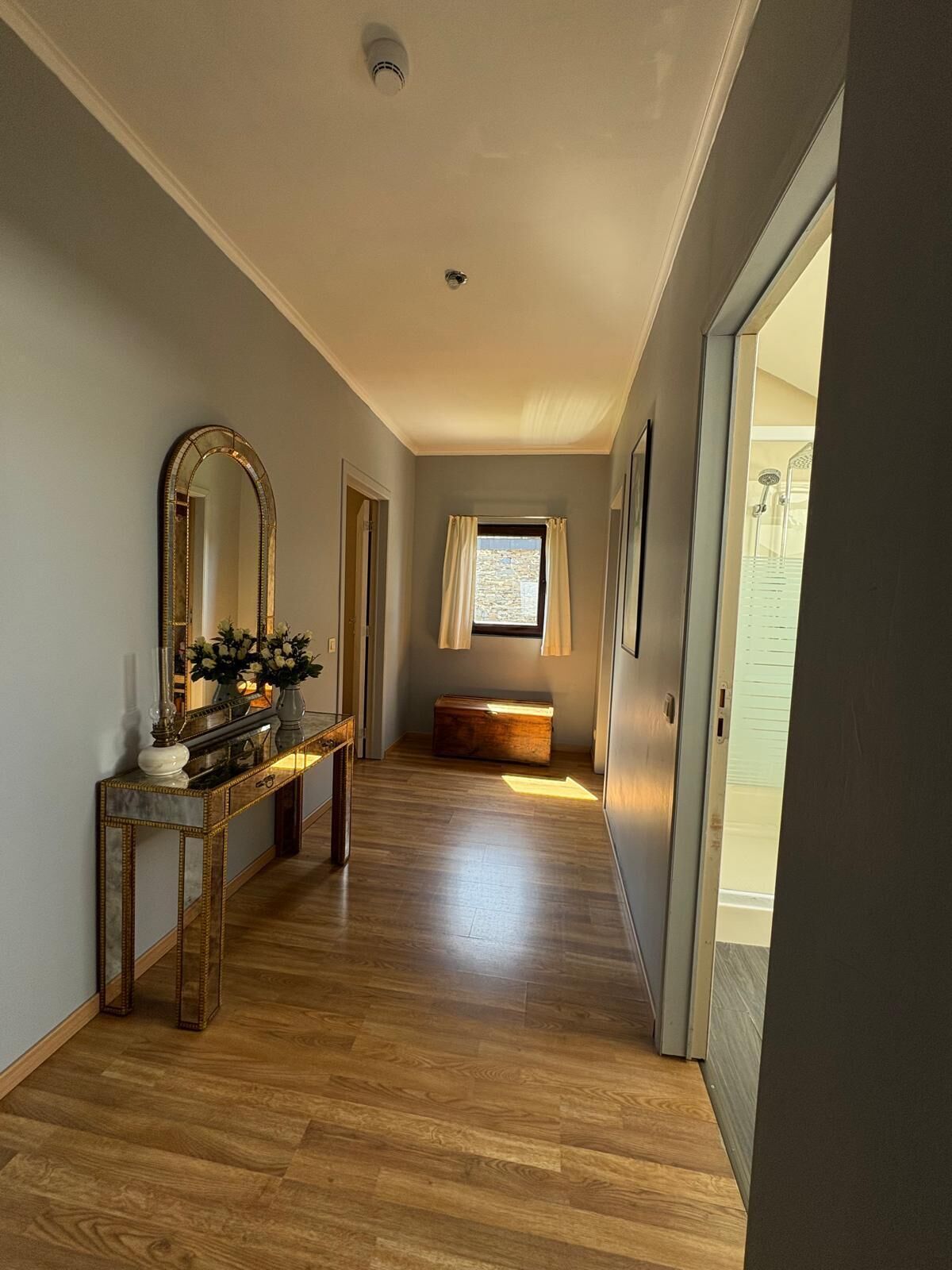 Un couloir doté d’un plancher en bois, une console avec un miroir et des fleurs, une fenêtre avec des rideaux et une porte donnant sur une salle de bain.