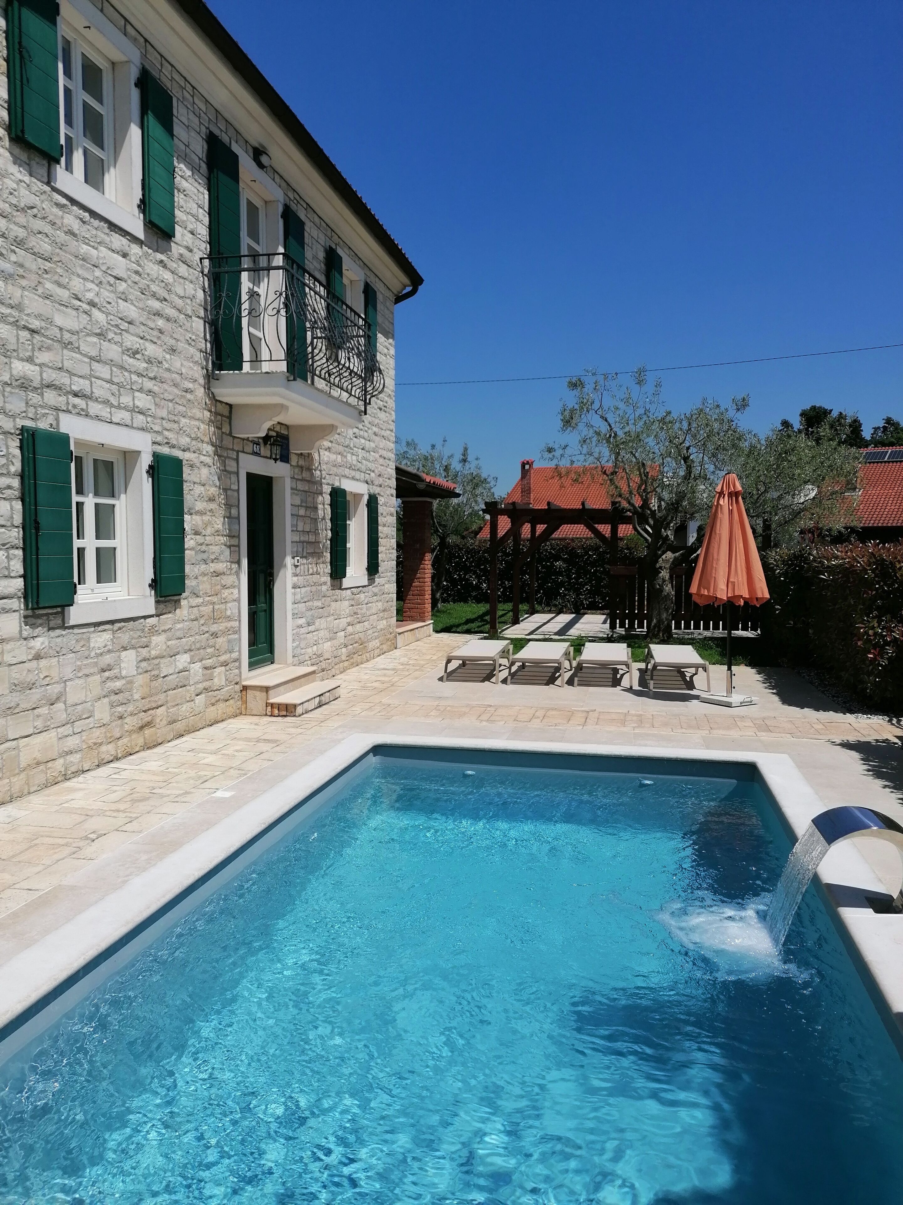 A swimming pool with a stone wall and a balcony with green shutters.