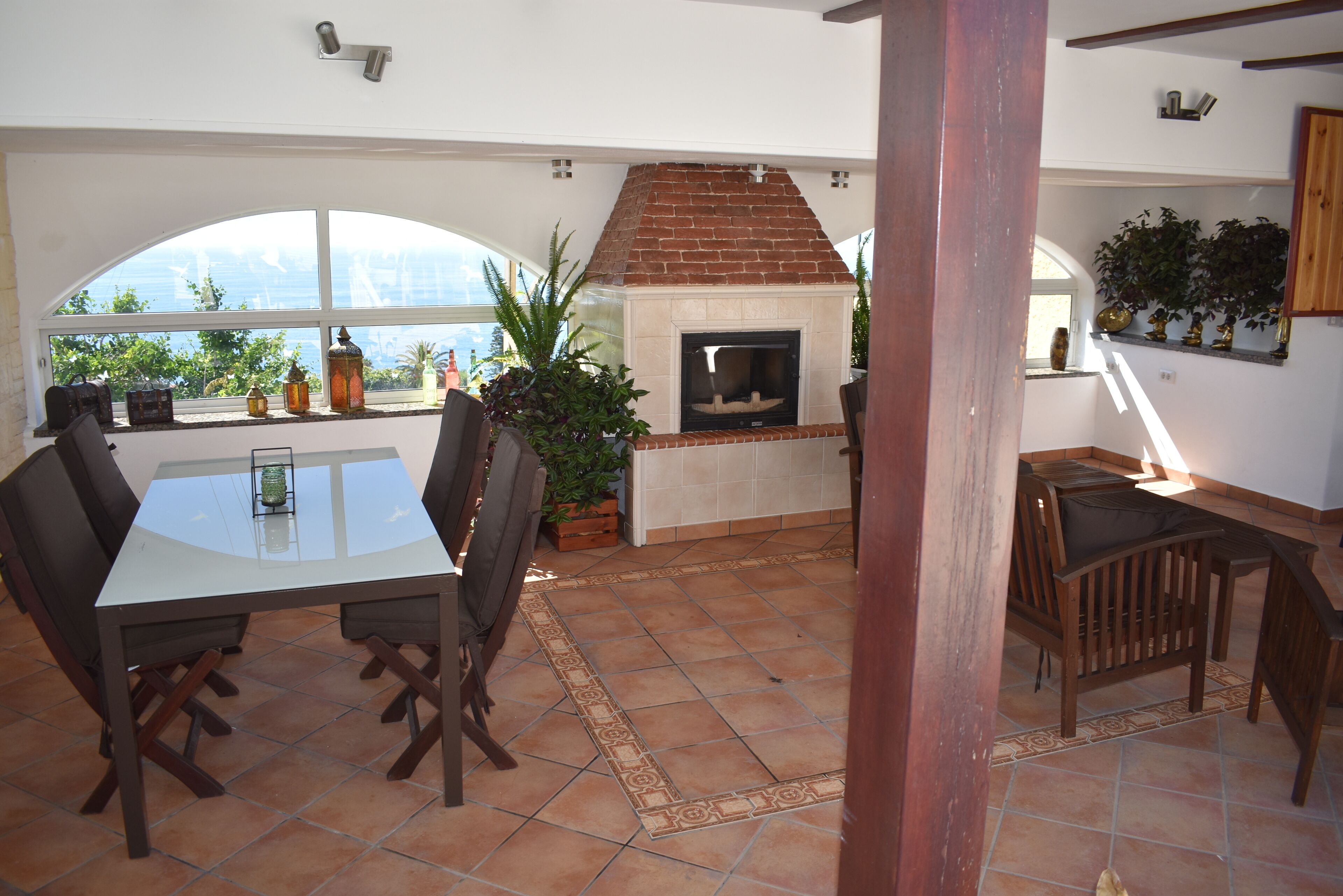 A dining area with a glass-top table, wooden chairs, a brick fireplace, and a view of the outdoors through large windows.