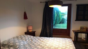 A bedroom with a bed, a nightstand, a lamp, a door with a view of greenery, and a small table with a picture frame. - Charming village house, Mt St Michel Bay (Genêts)
