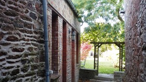 A stone building with a brick section, a metal pipe, and a stone pathway leading to a covered area with a bench. - Charming village house, Mt St Michel Bay (Genêts)