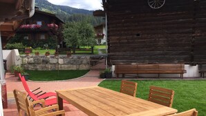 Ein Gartenmöbel-Set aus Holz mit Tisch und Stühlen auf einer Pflasterterrasse, mit einer Steinmauer und einem Holzgebäude im Hintergrund.