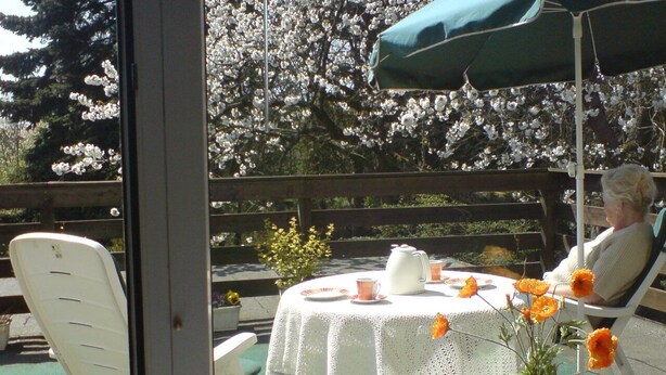 Une terrasse avec une table dressée pour deux personnes, une chaise et une vue sur un jardin où les fleurs sont en pleine floraison.
