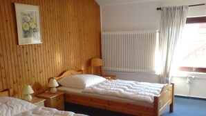 Une chambre avec deux lits en bois, une tête de lit en bois, une table de chevet avec des lampes, une fenêtre avec des stores et un tableau au mur.