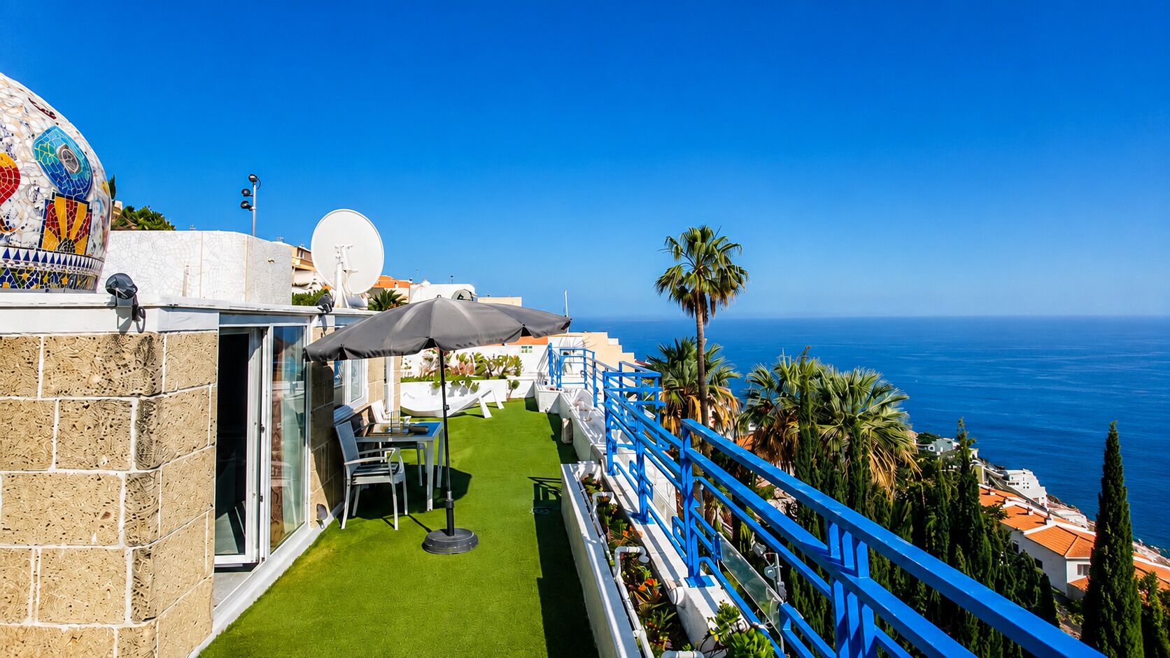 A balcony with a view of the sea, a table with chairs, and a blue railing.