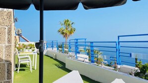Eine Dachterrasse mit weißen Liegestühlen, einem schwarzen Sonnenschirm und freiem Blick auf das Meer.