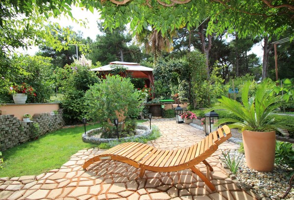 Une terrasse de jardin agrémentée d’un fauteuil de relaxation en bois, d’un chemin en pierre et de diverses plantes et arbres.