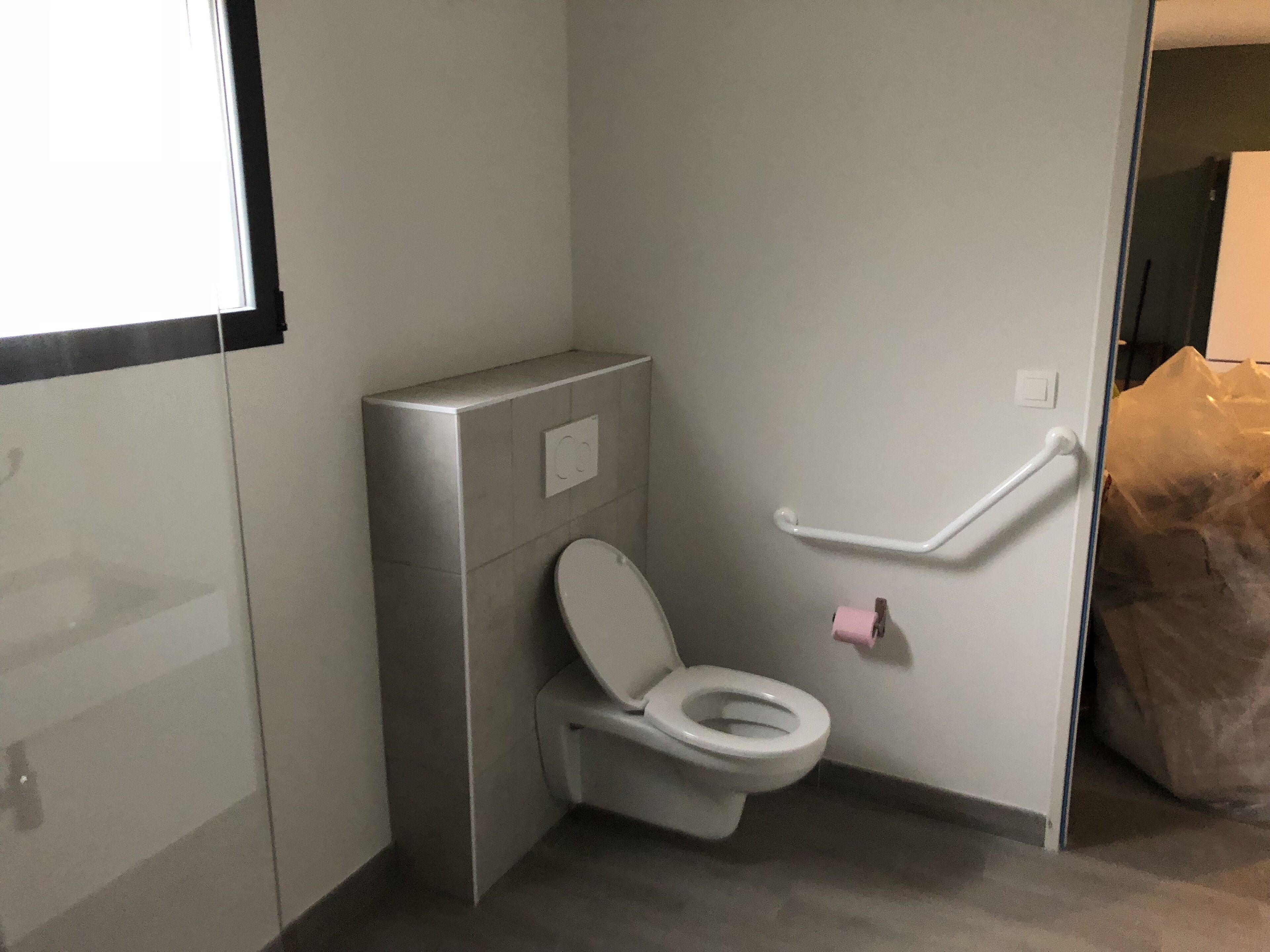 Ein kleines, modernes Badezimmer mit Wandtoilette, Handlauf und sichtbarem Mülleimer.
