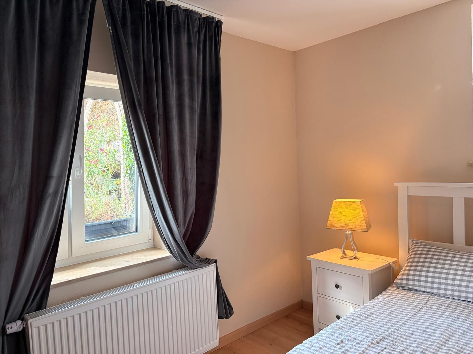 Un dormitorio con ventana, lámpara de noche, radiador y cama con edredón a cuadros.