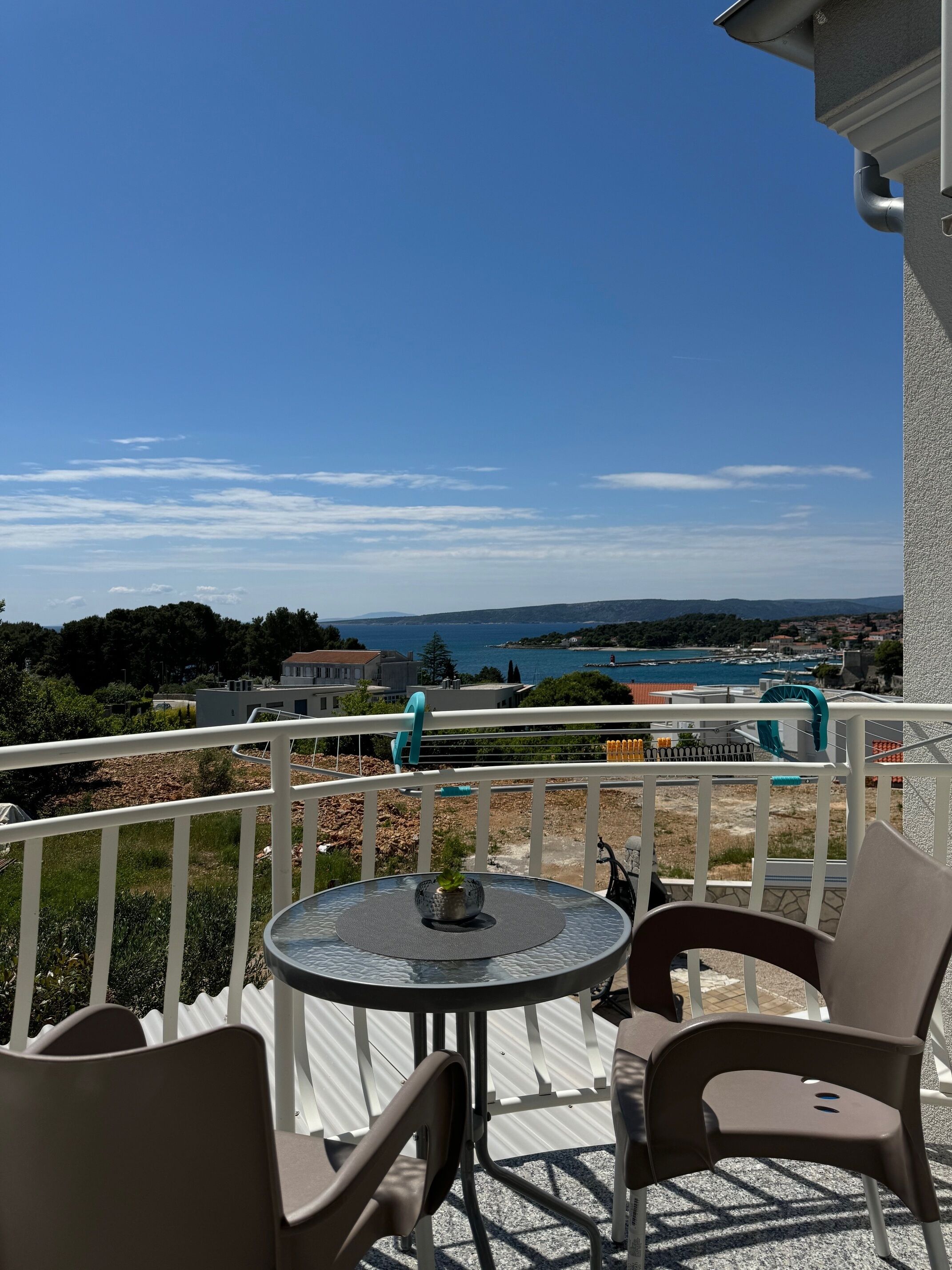 Ein Balkon mit rundem Tisch und zwei Stühlen, der Blick geht auf die Küste mit Häusern und einem Gewässer.