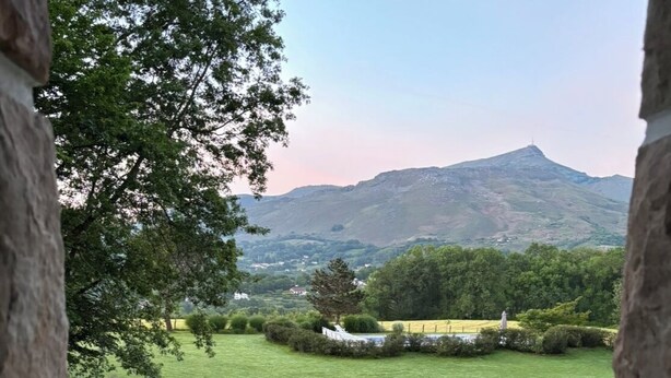 Vista desde una ventana enmarcada en piedra sobre un césped verde y exuberante, con rosas rosadas en primer plano, una montaña distante y un pequeño edificio blanco al fondo.