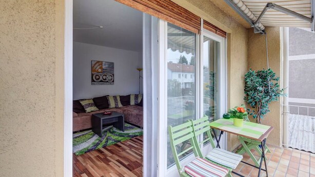 A balcony with a wooden floor, a green chair, a small table with a plant, and a sliding glass door leading to an interior space with a sofa and a coffee table.