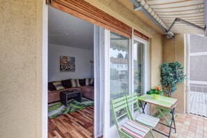 Uma varanda com piso de madeira, uma cadeira verde, uma pequena mesa com uma planta e uma porta de vidro deslizante que dá para um espaço interior com um sofá e uma mesa de centro.