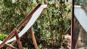 Una sedia di legno con seduta e schienale bianchi, posizionata all'aperto con vegetazione sullo sfondo.