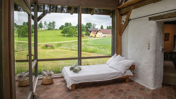 A bedroom with a large window, a bed, and a view of a house and fields.
