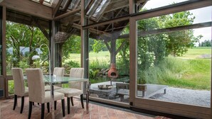 A glass-enclosed patio with a dining table and chairs, a fire pit, and a view of a grassy landscape.