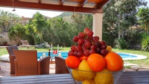 Uma tigela de vidro cheia de frutas sobre uma mesa de madeira ao ar livre, com uma piscina e espreguiçadeiras ao fundo.