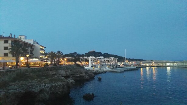 Uma vila costeira ao entardecer, com um porto de iates, edifícios e costa rochosa.