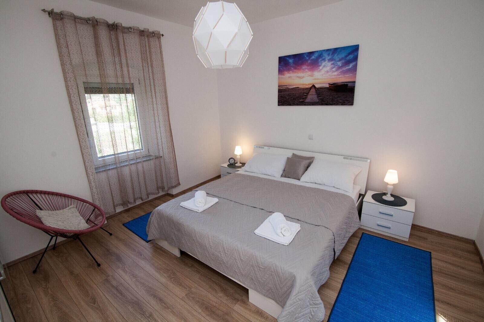 Ein Schlafzimmer mit einem Bett, zwei Nachttischen, einem Sessel, einem Fenster mit Vorhängen, einem Wandbild und einer Pendelleuchte.