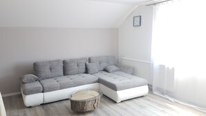 Un canapé d’angle moderne, de forme en L, avec des coussins gris et une table basse en verre dont le pied est constitué d’une souche d’arbre, placé dans une pièce lumineuse avec un radiateur blanc et une horloge encadrée accrochée au mur.