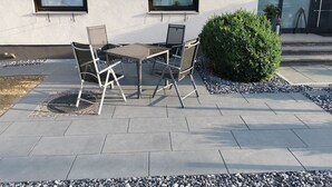 Une terrasse extérieure avec une table et des chaises, un petit arbuste et un bâtiment doté de deux fenêtres et d’une porte.