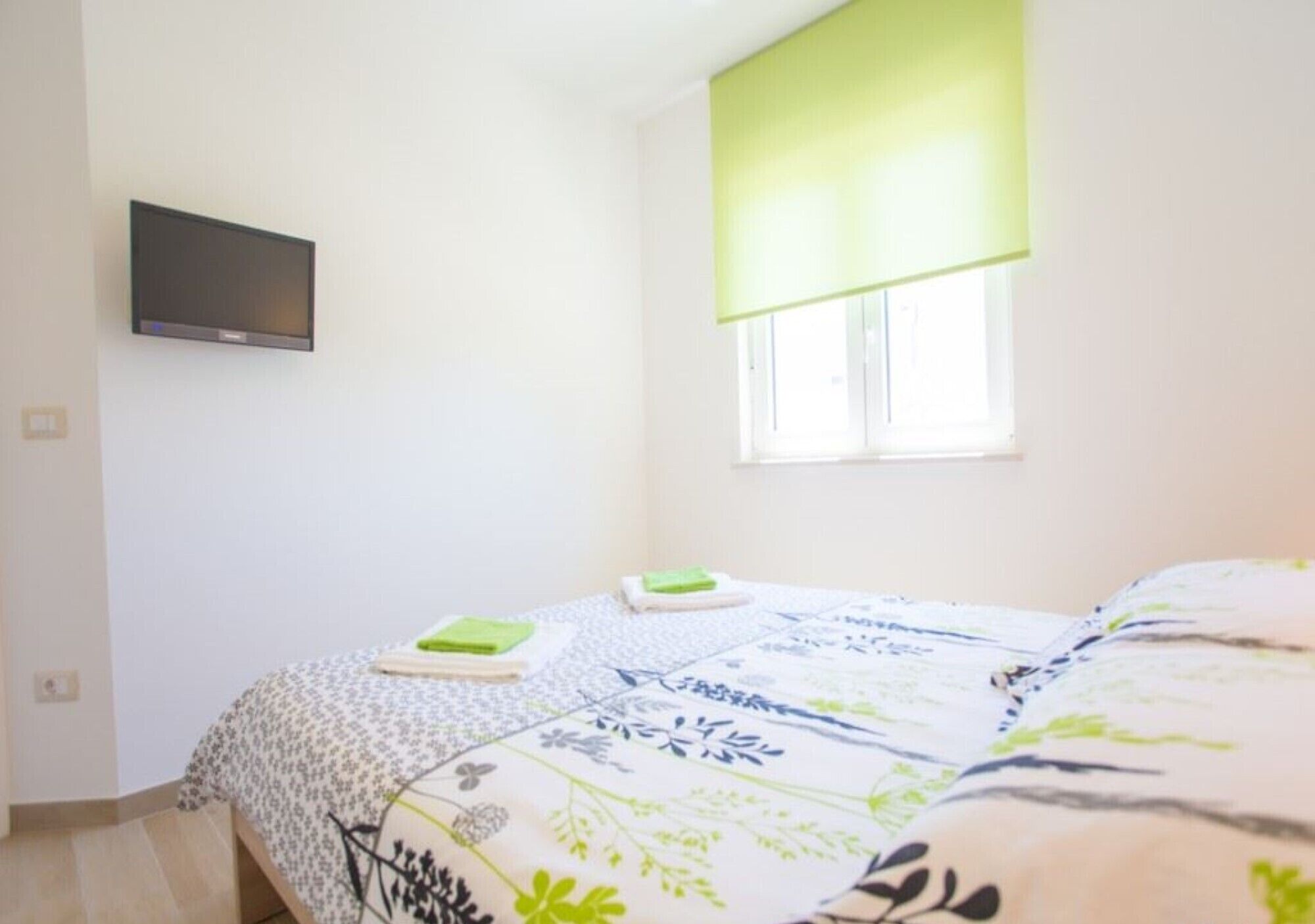 Een slaapkamer met een bed, een flatscreen televisie aan de muur, een raam met groene zonwering en een bed met een dessin.