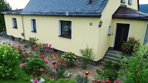 A yellow house with a tiled roof and a small garden with various flowers and shrubs.